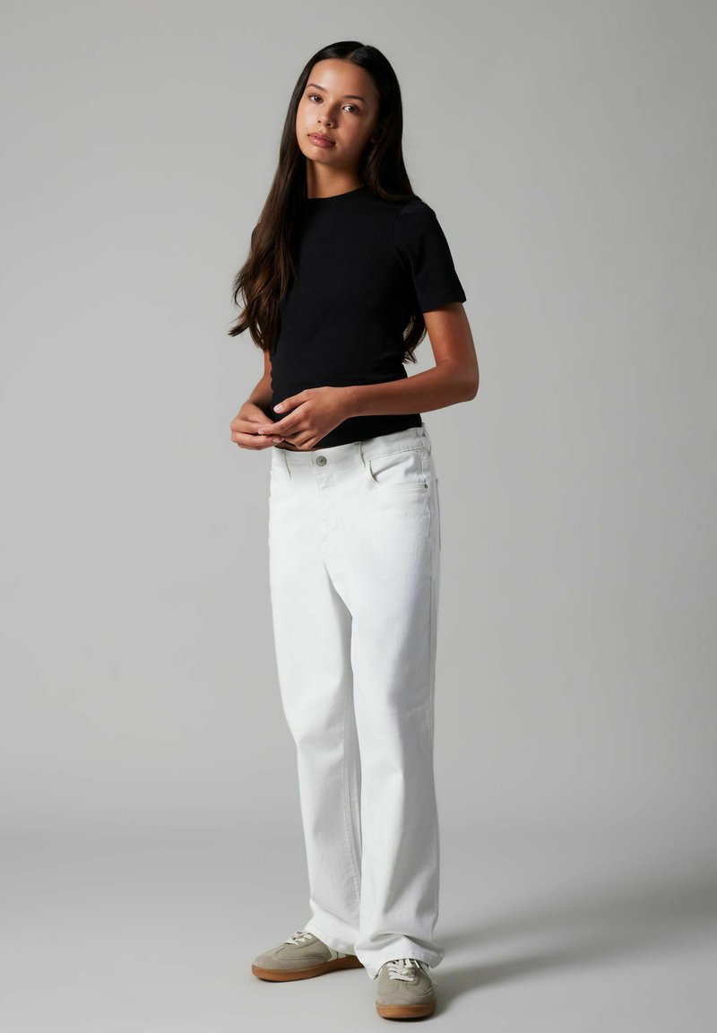 Jeune femme aux longs cheveux noirs, portant un haut noir à manches courtes, un jean blanc et des baskets beige, debout devant un fond gris uni.