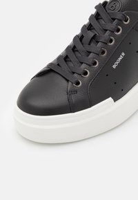 Baskets en cuir noir avec semelle blanche épaisse, lacets noirs, œillets métalliques, petites perforations et marquage "BOGNER" sur le côté.