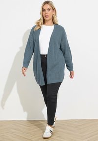 Un cardigan tricoté bleu clair avec une coupe décontractée, une ouverture à l'avant et des manches longues, associé à un pantalon noir et des baskets blanches.