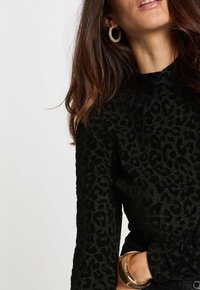 Donna che indossa un top nero con stampa leopardata, orecchini a cerchio dorati e un bracciale dorato spesso, con lunghi capelli castani mossi.