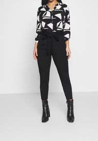 Černé kalhoty s vysokým pasem a zavázaným páskem, spárované s černo-bílou halenkou s abstraktním vzorem a černými kotníkovými botami.