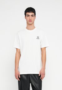 Balti medvilninė trumparankovė marškinėliai su mažu juodu grafiniu logotipu kairėje krūtinės pusėje. Dėvimi su juodomis odinėmis kelnėmis.