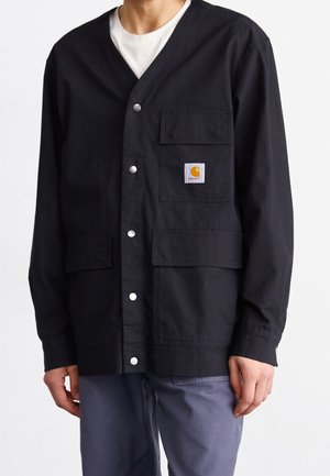Homme portant une veste de travail noire boutonnée avec trois poches avant et un petit patch carré avec logo sur la poche supérieure gauche.