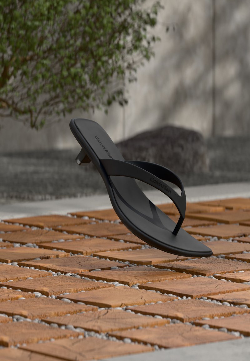 Μαύρο σανδάλι flip-flop Calvin Klein με μικρό τακούνι, τοποθετημένο σε ξύλινο πλακάκι σε εξωτερικό χώρο κοντά σε θολό τοίχο και φυτά.