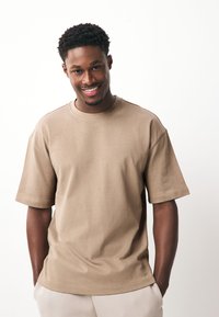 Pruun lühikeste varrukatega t-särk, valmistatud pehmest kangast, lõdva lõikega, ümar kaelusega ja lihtsate õmbluse detailidega. Kandmisel heledate värvidega püksid.