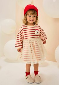 Rochie din pulover în dungi, cu talie elastică, straturi crem și roz, fustă din tull, căciulă de tricot roșie, șosete roșii până la genunchi și pantofi pufoși.