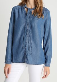 Donna che indossa una blusa in denim blu a maniche lunghe con scollatura a V e fiocco, abbinata a pantaloni bianchi, in piedi di fronte a uno sfondo neutro.