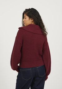 Bordowy sweter z dzianiny z szerokim kołnierzem, prążkowaną fakturą i balonowymi rękawami, w połączeniu z ciemnoniebieskimi dżinsami na jednolitym tle.