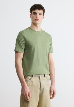 Jeune homme aux cheveux courts et bouclés portant un t-shirt à manches courtes vert clair et un pantalon beige sur un fond blanc uni.
