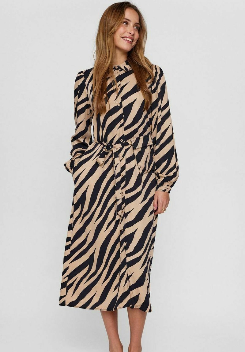 Rochie cu mâneci lungi, cu nasturi, de culoare bej cu dungi negre de zebra, având o talie legată și o siluetă evazată. Fabricată dintr-un material ușor.