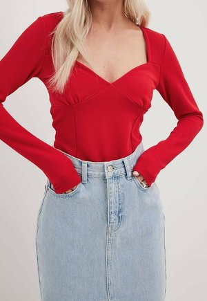 Woman wearing a fitted red long-sleeve top with a square neckline and light blue high-waisted denim skirt, hands in pockets.