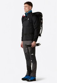 Svart fleecetröja med dragkedjeförsedda fickor, grå byxor med färgblockdesign och blå vandringsskor; man som håller i vandringsstavar.