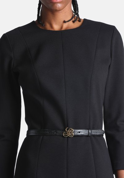 Personne portant une robe noire cintrée à manches longues et une fine ceinture noire avec une boucle décorative dorée à la taille.