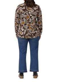 Frau trägt ein braunes, mit Paisleymuster versehenes Langarmoberteil, blaue Jeans mit rohem Saum und schwarze Schuhe. Sie steht mit dem Rücken zur Kamera vor hellem Hintergrund.