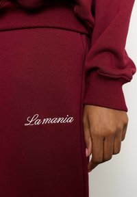 Burgundi spalvos megztinis su guma aptrauktomis rankovėmis ir balta siuvinėta etiketė "La mania" kairėje pusėje. Glotni tekstūra.