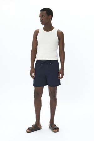 Weißes geripptes Tanktop, marineblaue Badeshorts mit Kordelzug und dunklen Sandalen. Schlichtes Design, keine Muster, leichtes Material.