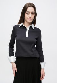 Čierna košeľa s dlhými rukávmi, bielym golierom a manžetami, so zapínaním na gombíky; nosená s čiernou plisovanou sukňou. Jednoduchý, čistý dizajn.