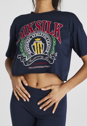 Vrouw draagt een marineblauwe crop top met rode tekst "SIKSILK" en een meerkleurig atletisch embleem, gecombineerd met marineblauwe high-waisted leggings.