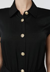 Robe-chemise noire sans manches avec col, ornée de cinq boutons dorés décoratifs alignés au centre devant.