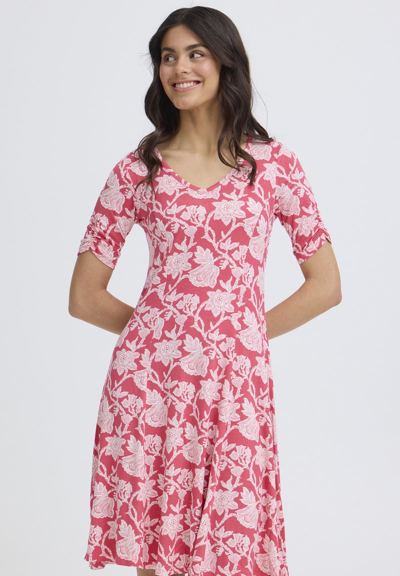 Femme souriante aux cheveux foncés portant une robe rose à motifs floraux, longueur genou et manches aux coudes, debout devant un fond clair uni.