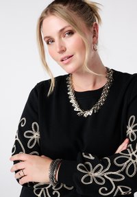 Zwarte sweatshirt met zilveren bloemenborduursels op de mouwen, gecombineerd met een zilveren statement ketting en gelaagde armbanden.