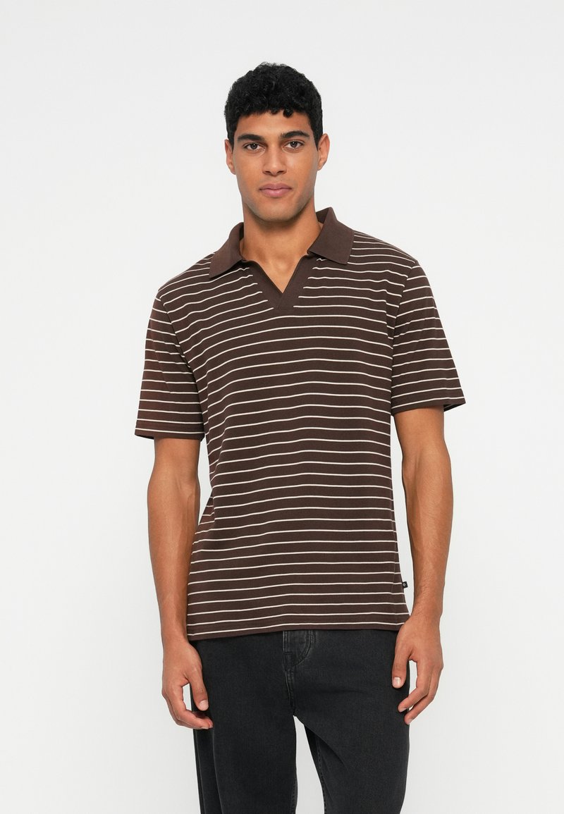 Man wearing a brown polo shirt with white horizontal stripes and black pants, standing against a plain light background.