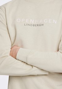 Felpa beige chiaro con collo a giro, caratterizzata dalla scritta "COPENHAGEN" e "LINDBERGH" stampate in colori a contrasto. Texture morbida e polsini aderenti.