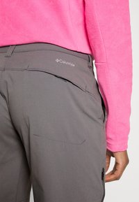 Pantalones grises de exterior con un logo en el bolsillo trasero, de textura suave y bolsillo lateral con cremallera. Combinados con una camiseta de fleece de color rosa brillante.