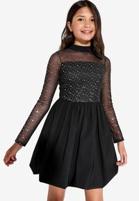 Schwarzes Kleid mit taillierter Oberseite, transparenten langen Ärmeln, die mit silbernen Punkten verziert sind, und einem plissierten Rock.