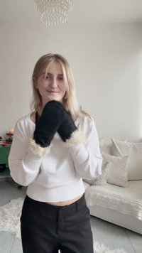 Fekete szőrme szegélyű kesztyű egy fehér pulóvert és fekete nadrágot viselő személyen, világos szobában, semleges tónusú bútorokkal.