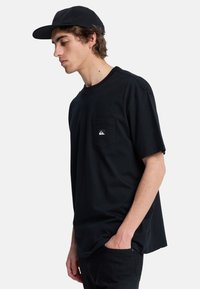 Sort bomulds t-shirt med korte ærmer og en brystlomme, der har et lille logo patch. Bæres med sorte bukser og en matchende kasket.