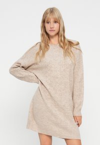 Ξανθιά γυναίκα που φοράει ένα μπεζ φόρεμα με πλέξη καλωδίου και μακριά μανίκια, ποζάρει με το ένα χέρι στη λεκάνη της απέναντι από έναν λευκό σκηνικό.