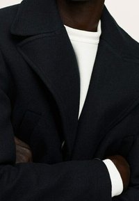 Czarny płaszcz wełniany z dużymi klapami i wyrazistymi ramionami, zestawiony z białym golfem. Szczegółami są boczne kieszenie i gładka tekstura.