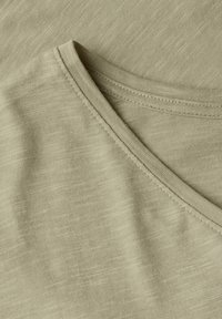 Béžová bavlněná tkanina s jemným texturovaným vzorem. Obsahuje výstřih do v a viditelné švy podél okrajů.