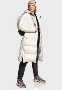 Lang, glanzende witte pufferjas met een capuchon, zijritsen en een zwarte binnenkant, gecombineerd met donkere broeken en grijs-bruin sneakers.