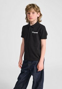 Svart bomullspikétröja med krage och tre knappar, med vitt "Hummel"-logotyp. Matchas med mörkblå denimbyxor.