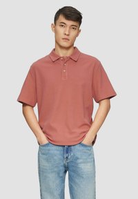 Jonge man met kort donkerhaar, in een stofroze polo met korte mouwen en lichtblauwe jeans, met zijn handen in zijn zakken.