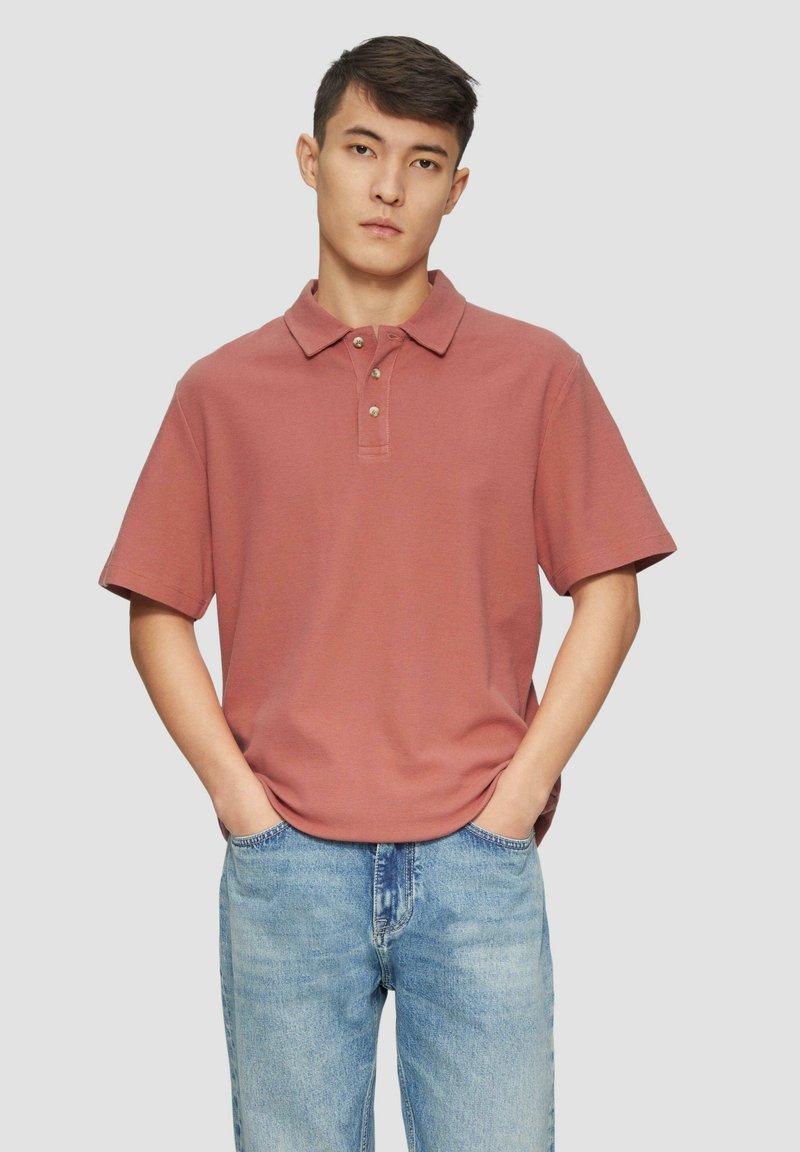 Jonge man met kort donkerhaar, in een stofroze polo met korte mouwen en lichtblauwe jeans, met zijn handen in zijn zakken.