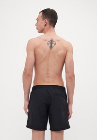 Pantaloni da nuoto neri con cintura elastica e una tasca sul retro. Un tatuaggio visibile di un fleur-de-lis si trova sulla schiena della persona.