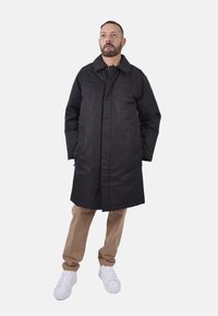 Cappotto nero, lungo fino al ginocchio, con colletto e tasche frontali; tessuto liscio; abbinato a pantaloni beige e sneakers bianche.