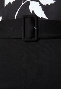 Svart tygbälte med rektangulär spänne, sex hål för justering, strukturerad yta, i kombination med ett slätt svart plagg nedanför.