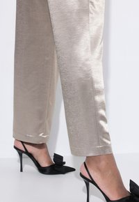 Donna che indossa pantaloni satin beige e décolleté neri con tacco alto e punte affusolate, decorati con fiocchi.