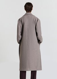 Lungo cappotto grigio in lana con collo a lancia, vestibilità comoda e dettagli di cuciture visibili. Il tessuto ha una texture morbida e un peso consistente.