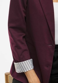 Persoană îmbrăcată cu un blazer vișiniu închis, cu manșete cu dungi albe pliate, purtând pantaloni negri și un top alb, așezată pe un scaun din lemn.