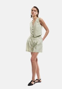 Lichtgroene linnen twee-delige outfit met een mouwloze knopenvest en geplooide shorts, gecombineerd met zwarte geweven sandalen.