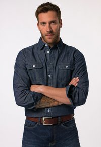 Camisa de mezclilla oscura con dos bolsillos en el pecho, mangas enrolladas y cuello tipo camisa, combinada con unos jeans oscuros y un cinturón de cuero marrón.