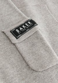 Sivá tkanina s textúrovaným povrchom, na ktorej je čierna nálepka s nápisom "BAKER by TED BAKER," umiestnená na oblasti vrecka.