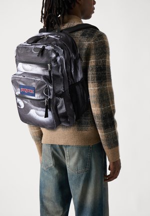 Black and gray Jansport backpack with multiple zippered compartments, mesh side pocket, and a textured surface, worn over a patterned sweater.