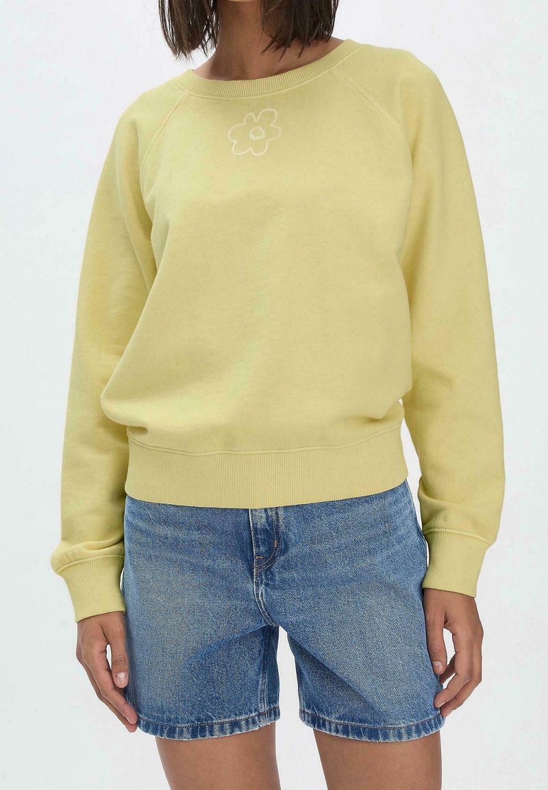 Personne portant un sweat-shirt jaune clair avec un petit motif floral associé à un short en denim bleu moyen, debout devant un fond uni.