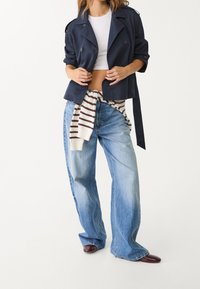 Marineblauwe trenchcoat over een wit cropped topje, gecombineerd met lichtblauwe loszittende spijkerbroek en een gestreepte trui geknoopt rond de taille. Bruine schoenen.